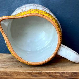 May include: A ceramic pitcher with a speckled white body and a curved handle. The rim is trimmed with a yellow and brown design. The interior is a light blue color.