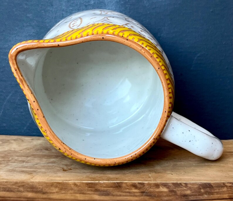 May include: A ceramic pitcher with a speckled white body and a curved handle. The rim is trimmed with a yellow and brown design. The interior is a light blue color.