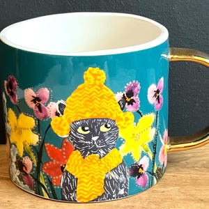 May include: Teal ceramic mug with a gold handle and rim. The mug features a black cat wearing a yellow hat and scarf, surrounded by colourful flowers. The interior is white.
