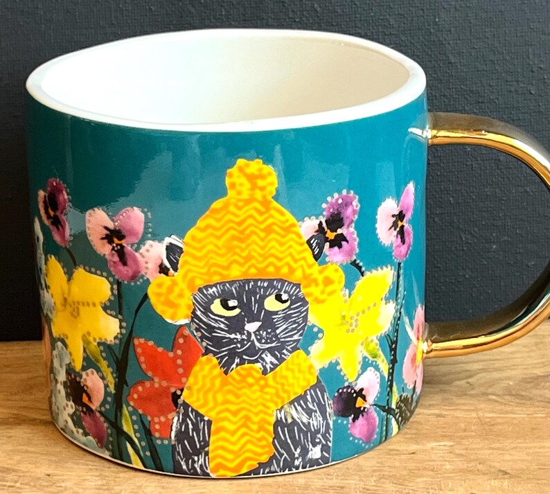 May include: Teal ceramic mug with a gold handle and rim. The mug features a black cat wearing a yellow hat and scarf, surrounded by colourful flowers. The interior is white.