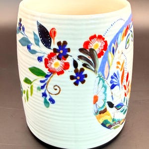 May include: White ceramic mug with a floral design. The mug features red, blue, and yellow flowers with green leaves. The mug has a ribbed texture and a black base.