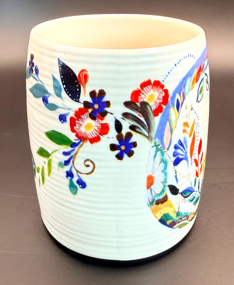 May include: White ceramic mug with a floral design. The mug features red, blue, and yellow flowers with green leaves. The mug has a ribbed texture and a black base.