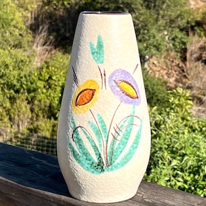 May include: Cream-colored ceramic vase with a floral design. The vase features yellow, orange, and purple flowers with green leaves and stems. The vase has a textured surface and a slightly flared shape.