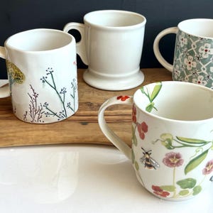 Pu&ograve; includere: Quattro tazze in ceramica bianca con manici sono esposte su un tagliere di legno. Una &egrave; semplice, una ha un motivo floreale e due sono decorate con disegni botanici. Le tazze variano in forma e dimensione.