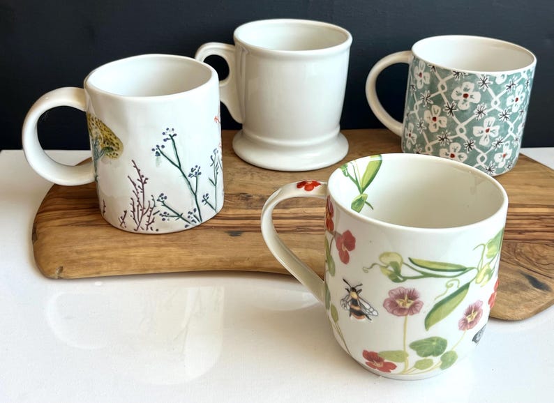 Pu&ograve; includere: Quattro tazze in ceramica bianca con manici sono esposte su un tagliere di legno. Una &egrave; semplice, una ha un motivo floreale e due sono decorate con disegni botanici. Le tazze variano in forma e dimensione.