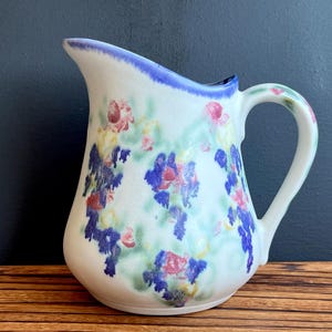 May include: White ceramic pitcher with a blue rim and handle, adorned with a floral pattern in shades of blue, pink, and yellow. The pitcher has a curved spout and a rounded body, suitable for serving beverages.