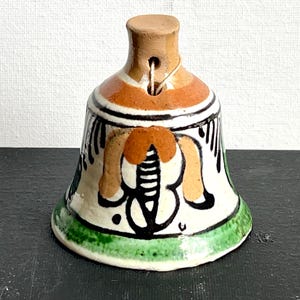 May include: A small, hand-painted ceramic bell with a green, orange, and black floral design. The bell has a brown clay top and a string attached to the top.