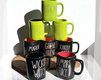 May include: A collection of Halloween-themed mugs in various colors. The mugs are black and lime green, with phrases like "Potion", "Boo to You", and "Wicked Witch" written on them. The mugs are stacked on a wooden surface.