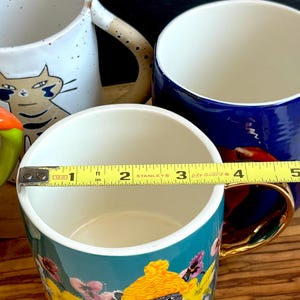 May include: A selection of ceramic mugs with different patterns. One mug has a cat design, another is plain blue, and a third is teal with a floral and cat motif. A measuring tape indicates the mug's diameter is about 7.6 cm.