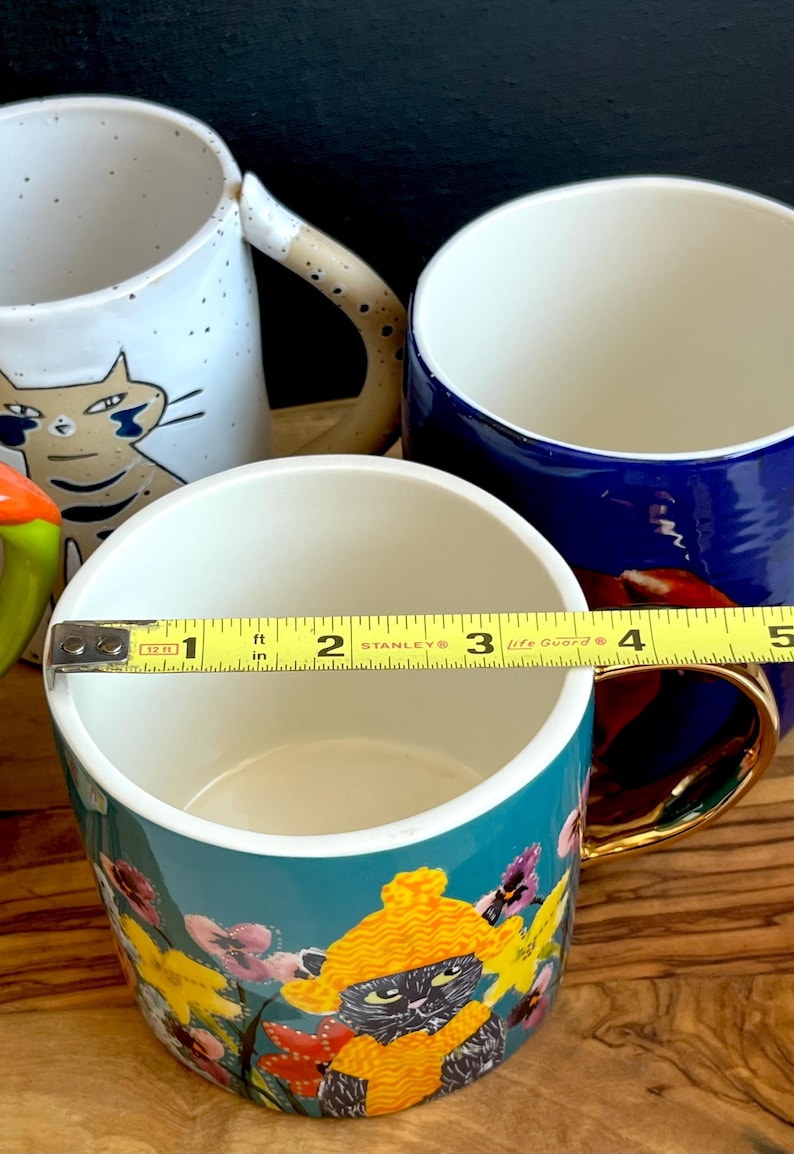 May include: A selection of ceramic mugs with different patterns. One mug has a cat design, another is plain blue, and a third is teal with a floral and cat motif. A measuring tape indicates the mug's diameter is about 7.6 cm.