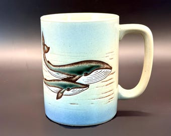 May include: A ceramic mug with a light blue gradient and a whale design. The whales are teal and brown, with white detailing. The mug has a curved handle and is suitable for hot or cold beverages.