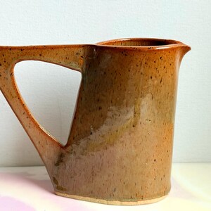 May include: A ceramic pitcher with a glossy, speckled tan and brown glaze. The vessel features a wide, angular handle that connects to the body at the top and bottom, creating a triangular negative space. It has a simple, flared pouring spout.