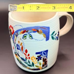 May include: White ceramic mug with a floral design and a light pink handle. The mug is approximately 3 inches (7.6 cm) in diameter and features a colorful, hand-painted floral pattern in blue, red, and green.
