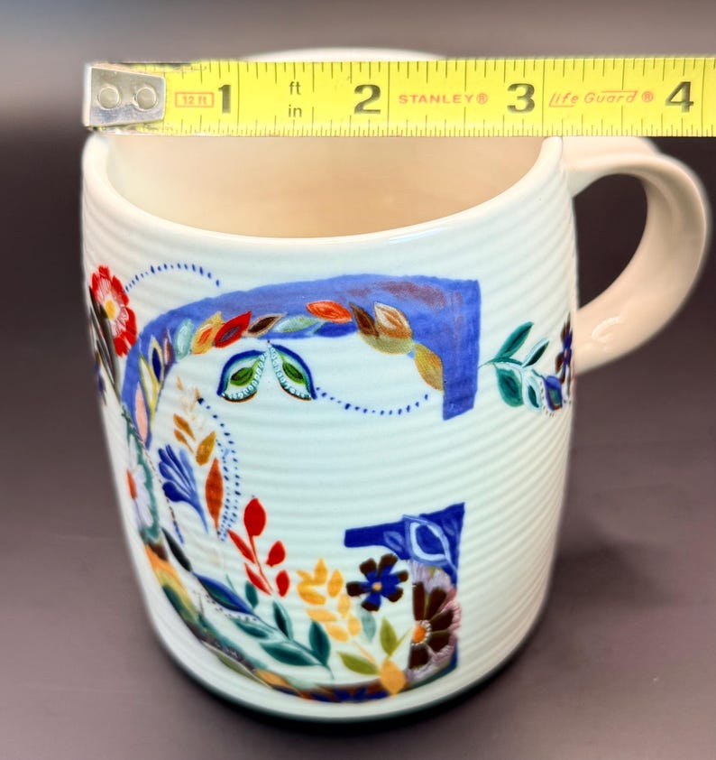 May include: White ceramic mug with a floral design and a light pink handle. The mug is approximately 3 inches (7.6 cm) in diameter and features a colorful, hand-painted floral pattern in blue, red, and green.