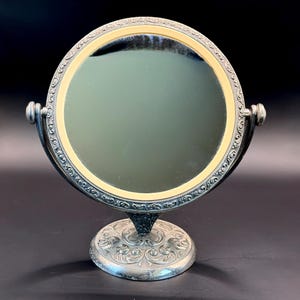 May include: A silver-toned vanity mirror with a round, pivoting mirror. The mirror has a cream-colored border and ornate detailing around the edge. The base is also silver-toned with a decorative floral design.