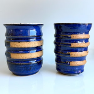 May include: Two handmade ceramic vases. The vases are a deep blue with horizontal bands of tan. The vases have a textured, ribbed design. The rims of the vases are white. The vases are displayed on a white surface.