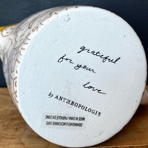 May include: White ceramic pitcher with a yellow handle and floral design. The bottom of the pitcher has the words "grateful for your love" written in black script, along with the brand name "by ANTHROPOLOGIE".
