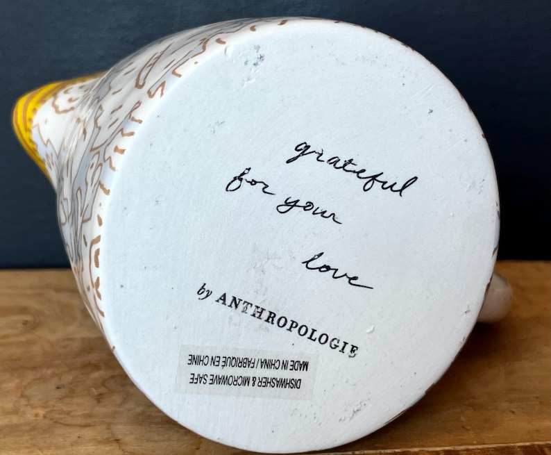 May include: White ceramic pitcher with a yellow handle and floral design. The bottom of the pitcher has the words "grateful for your love" written in black script, along with the brand name "by ANTHROPOLOGIE".