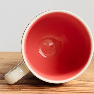 May include: A white ceramic mug with a red interior. The mug has a rounded handle.