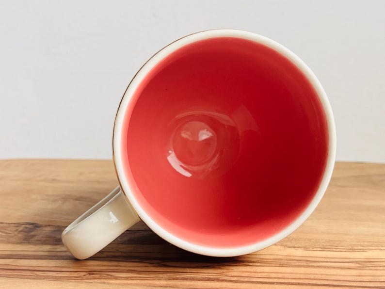 May include: A white ceramic mug with a red interior. The mug has a rounded handle.