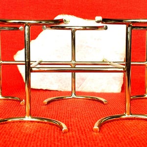 May include: A set of three gold-coloured metal display stands. Each stand features a curved base, vertical supports, and a horizontal bar. The stands are arranged on a red textured surface.