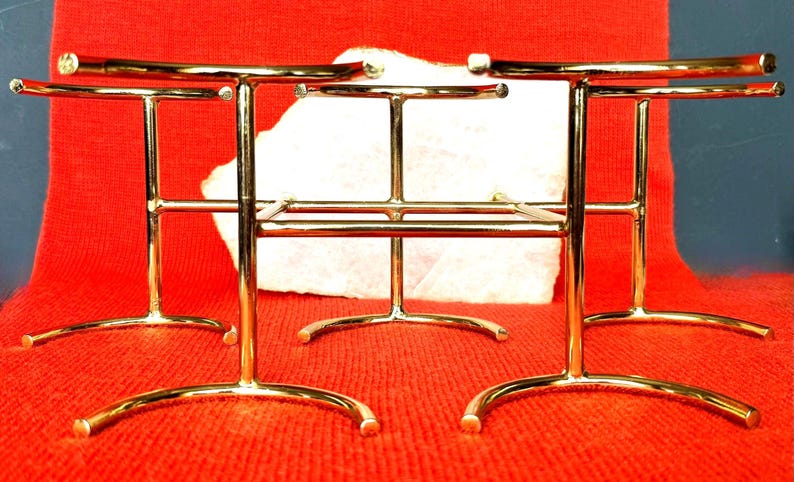 May include: A set of three gold-coloured metal display stands. Each stand features a curved base, vertical supports, and a horizontal bar. The stands are arranged on a red textured surface.