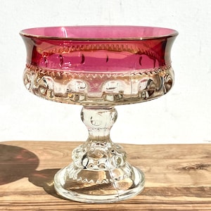 May include: A vintage glass pedestal bowl with a clear base and a deep red, ruby-colored top. The bowl has a decorative, scalloped rim and a textured, patterned base.