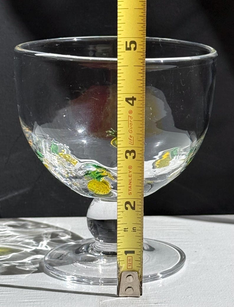 May include: A clear glass goblet with a wide bowl and a short stem. The goblet is decorated with yellow and green pineapple designs around the rim. The height of the goblet is approximately 5 inches.