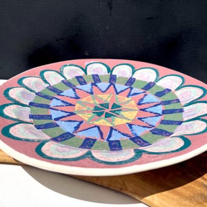 May include: A round, hand-painted ceramic plate with a pink base and a colorful geometric design. The design features a central starburst pattern in shades of yellow, pink, and green, surrounded by blue and green shapes. The plate is approximately 10 inches in diameter.