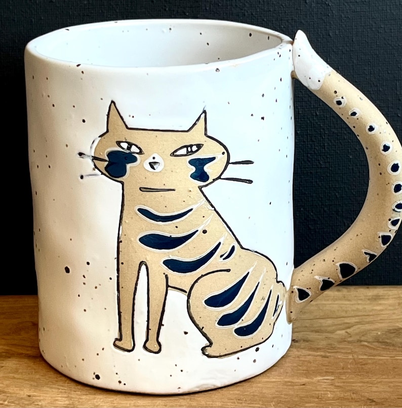 May include: A white ceramic mug featuring a hand-painted cat design. The cat is tan with blue accents and black whiskers. The handle is tail-shaped with black spots. The mug has a speckled finish.