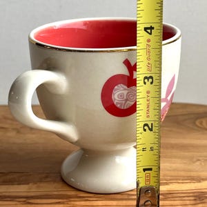 May include: A white ceramic mug with a red and pink apple design. The mug has a gold rim and a pedestal base. The mug is 3.5 inches tall.