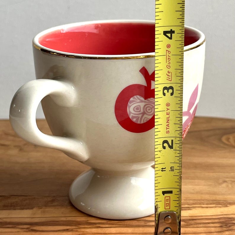 May include: A white ceramic mug with a red and pink apple design. The mug has a gold rim and a pedestal base. The mug is 3.5 inches tall.
