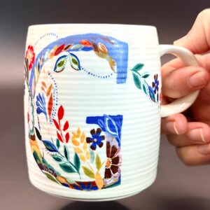 May include: White ceramic mug with a large, colorful floral "G" design. The letter is painted with blue, red, orange, and green leaves and flowers. The mug has a ribbed texture and a white handle.