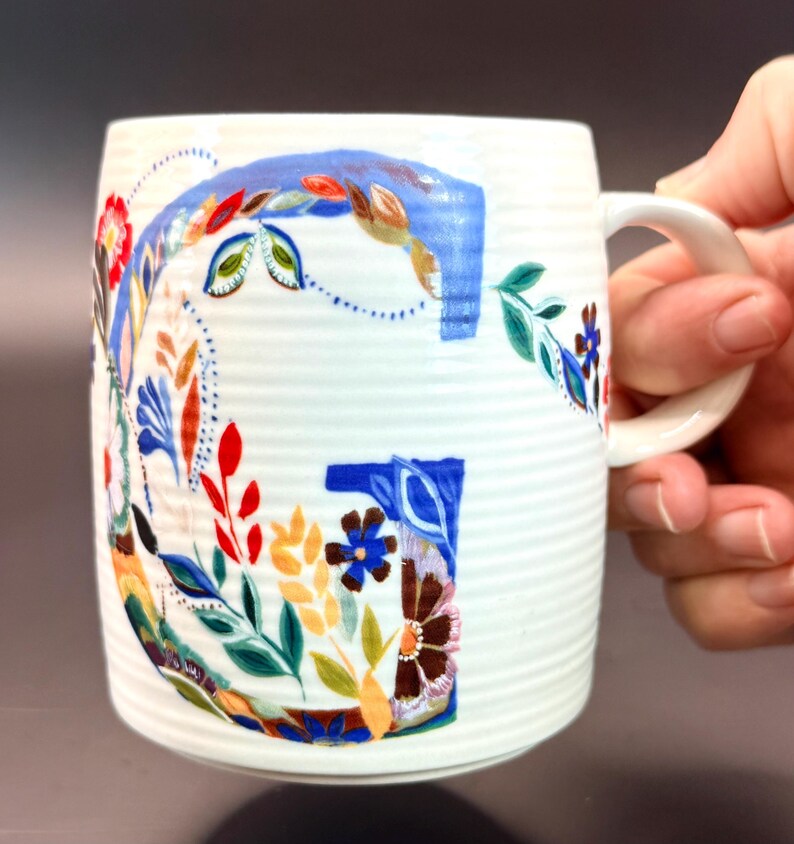 May include: White ceramic mug with a large, colorful floral "G" design. The letter is painted with blue, red, orange, and green leaves and flowers. The mug has a ribbed texture and a white handle.