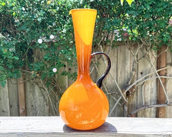 May include: A tall, orange glass pitcher with a black handle. The pitcher has a rounded base and a slender neck that flares out at the top. The handle is curved and attached to the side of the pitcher. The pitcher is sitting on a wooden surface.