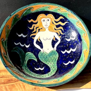 May include: A ceramic bowl with a mermaid design. The mermaid has long, flowing red hair and a green tail. The bowl is decorated with a blue background and white waves. The rim of the bowl is decorated with a yellow and green border.