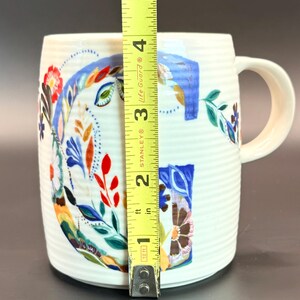 May include: White ceramic mug with a floral design and the letter "G". The mug is approximately 4 inches tall. The floral design includes red, blue, and brown elements. The mug has a ribbed texture and a curved handle.