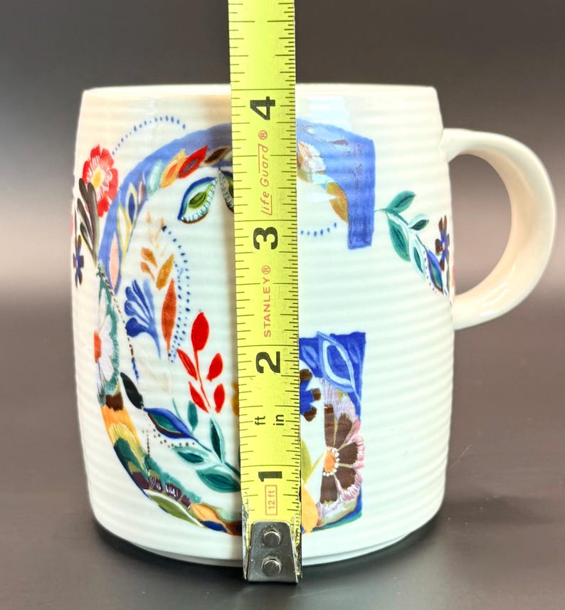 May include: White ceramic mug with a floral design and the letter "G". The mug is approximately 4 inches tall. The floral design includes red, blue, and brown elements. The mug has a ribbed texture and a curved handle.