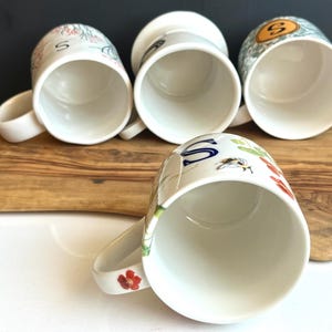 Pu&ograve; includere: Quattro tazze in ceramica bianca con manici, ognuna con disegni floreali e di lettere unici. Una tazza mostra una grande "S" blu, mentre altre presentano illustrazioni botaniche colorate. Le tazze sono disposte su una superficie di legno.