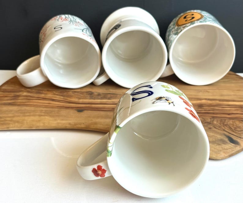 Pu&ograve; includere: Quattro tazze in ceramica bianca con manici, ognuna con disegni floreali e di lettere unici. Una tazza mostra una grande "S" blu, mentre altre presentano illustrazioni botaniche colorate. Le tazze sono disposte su una superficie di legno.