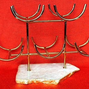 May include: A gold-coloured metal wine glass rack with six curved holders. The rack is mounted on a white and gold-edged stone base. The background is a solid red colour.