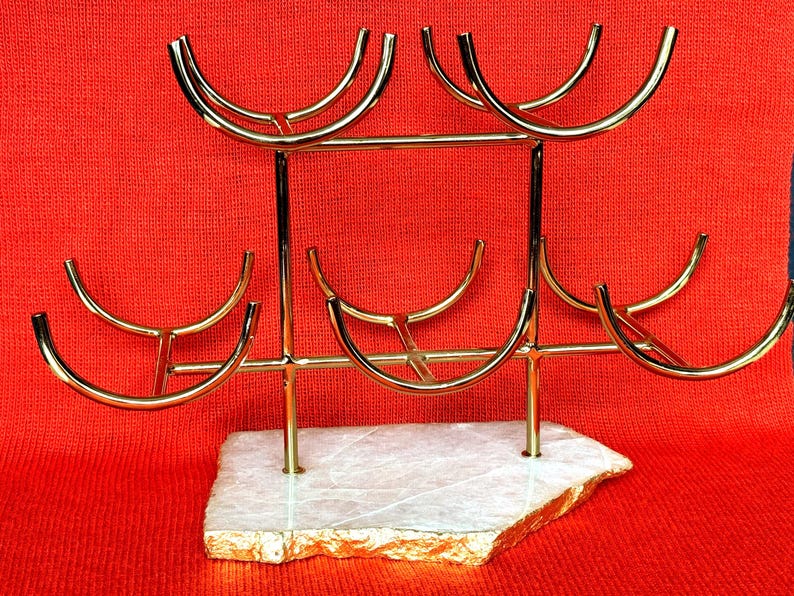 May include: A gold-coloured metal wine glass rack with six curved holders. The rack is mounted on a white and gold-edged stone base. The background is a solid red colour.