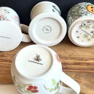 Pu&ograve; includere: Una collezione di tazze in ceramica bianca con disegni floreali e botanici. Le tazze presentano vari motivi, tra cui fiori, foglie e un'ape. I fondi delle tazze mostrano il logo del marchio "Anthropologie". Alcune tazze hanno la lettera "S" o un disegno di uccello.
