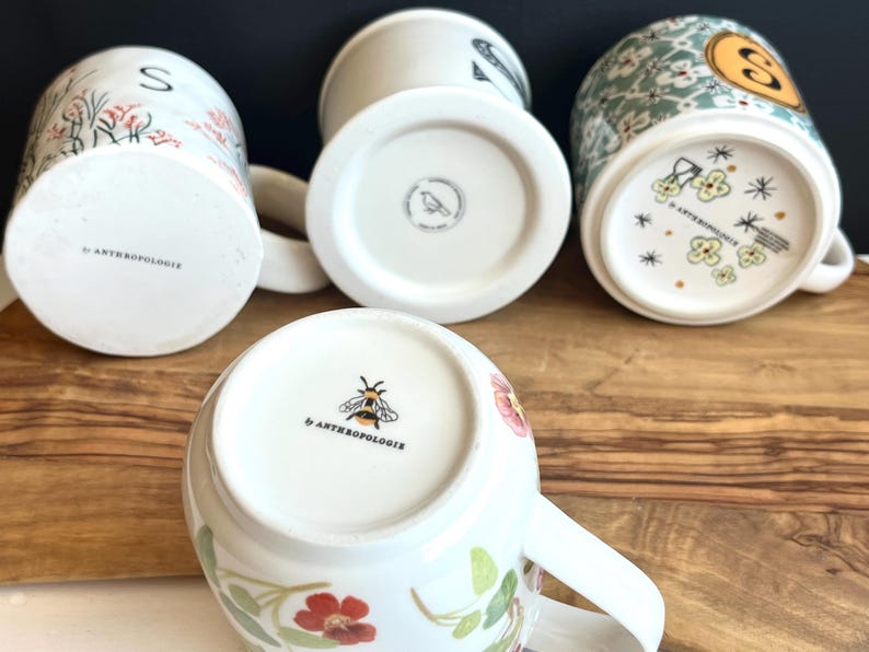 Pu&ograve; includere: Una collezione di tazze in ceramica bianca con disegni floreali e botanici. Le tazze presentano vari motivi, tra cui fiori, foglie e un'ape. I fondi delle tazze mostrano il logo del marchio "Anthropologie". Alcune tazze hanno la lettera "S" o un disegno di uccello.