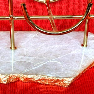 May include: A gold-coloured metal decorative stand with a white stone base. The stone has gold-coloured edges and a natural, textured appearance. The stand is set against a red background.