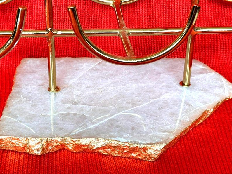 May include: A gold-coloured metal decorative stand with a white stone base. The stone has gold-coloured edges and a natural, textured appearance. The stand is set against a red background.