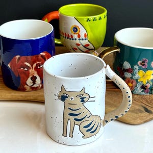 May include: Four ceramic mugs with distinctive designs. One mug displays a dog portrait on a blue backdrop. Another has a cat illustration. The other two mugs feature floral and abstract patterns. Each mug has a differently coloured handle.