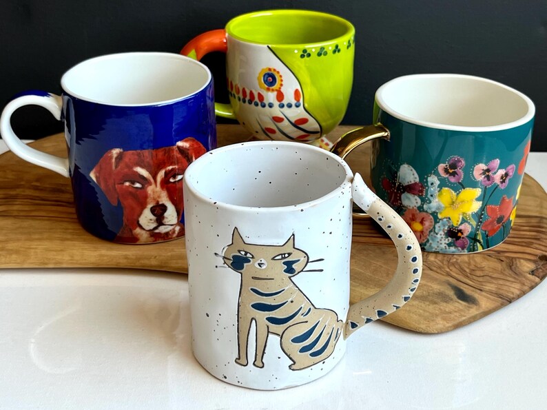 May include: Four ceramic mugs with distinctive designs. One mug displays a dog portrait on a blue backdrop. Another has a cat illustration. The other two mugs feature floral and abstract patterns. Each mug has a differently coloured handle.