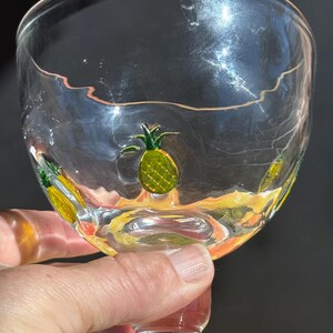 May include: Clear glass goblet with a wide bowl and a red stem. The glass is decorated with yellow and green pineapple designs around the bowl. The goblet is held by a hand.