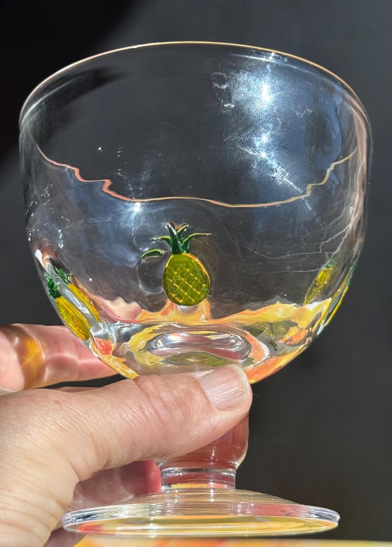 May include: Clear glass goblet with a wide bowl and a red stem. The glass is decorated with yellow and green pineapple designs around the bowl. The goblet is held by a hand.